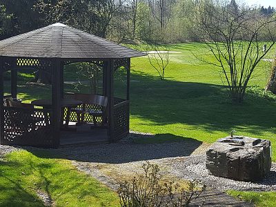 A wooden gazebo with a peaked roof stands beside a circular stone fire pit amidst a green lawn, surrounded by trees, under a sunny sky.