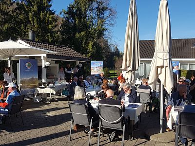 People sit at tables on a sunny patio, surrounded by trees and umbrellas, enjoying an outdoor event.