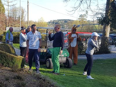 People at a golf event, some talking, one in a costume, greenery and a golf cart visible, creating a social and playful atmosphere.
