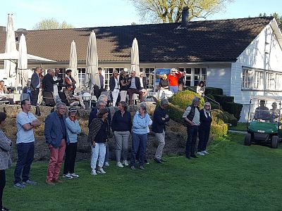 A group of people standing and sitting outside a golf club under umbrellas, watching a golf game. The setting is sunny with a clear blue sky.