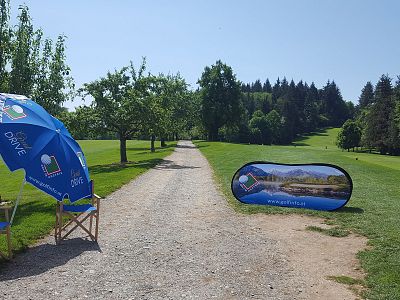 A golf course with a gravel path surrounded by trees and grass. Blue promotional umbrellas and signs are on the left. Clear blue sky overhead.