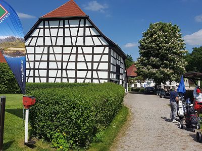 Traditional half-timbered house with red roof; paved path, manicured hedges, tree in bloom, and people with golf bags on sunny day.