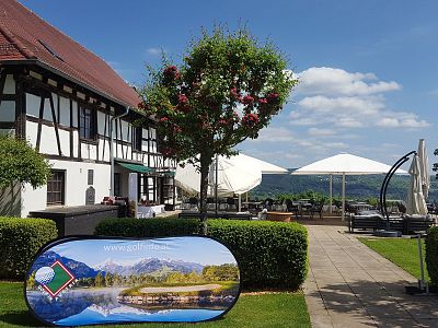 Countryside scene with a half-timbered house, garden, umbrellas, and distant hills under a blue sky. A landscape banner is also visible.