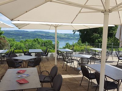 An outdoor terrace with tables and chairs under umbrellas, overlooking a lush green landscape with a lake and hills in the background.