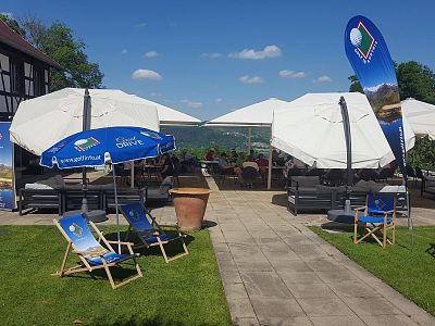 An outdoor area with white umbrellas and blue deck chairs on a patio. There's a scenic view with green grass, a clear sky, and promotional flags.