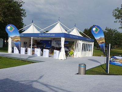White outdoor tent pavilion displaying Salzburgerland banners, set up on a sunny day with clear skies, surrounded by grass and trees.
