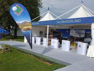 Outdoor event booth with Salzburgerland banners, brochures on tables, and seating. Tents and green grass are visible under a clear sky.