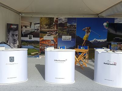 Exhibition booth with white stands displaying brochures, travel destination images, and logos, set in a tent-like structure.