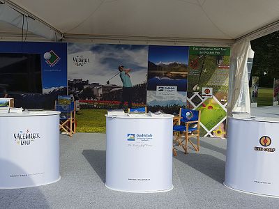 Outdoor booth with three white display counters in front of a golfing backdrop. The booth is part of a golf-themed event with promotional materials.