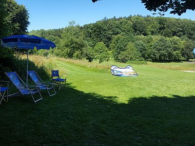 A sunny meadow with blue chairs and umbrellas under trees, lush grass, and a forest in the background, creating a peaceful retreat setting.