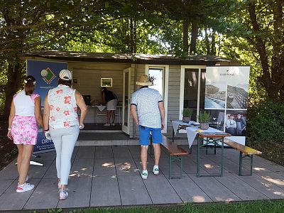 Three people walking towards a small wooden shed surrounded by trees, with a table and bench outside under a shaded area.