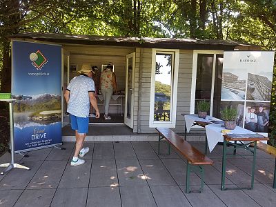 Outdoor booth with two people, surrounded by trees. One enters, the other is inside. Banners and table with pamphlets are visible.