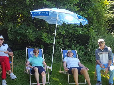 Four people sitting in deck chairs under a blue umbrella, surrounded by trees and grass, enjoying a sunny day.