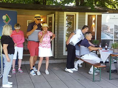 People at a golf event, casually dressed, near a wooden cabin with banners. Some stand while others sit at a bench, enjoying the outdoor setting.