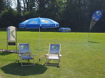 Two deck chairs with a blue umbrella on a grassy field, surrounded by advertising banners. Trees and clear sky in the background create an outdoor event vibe.