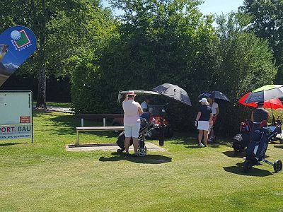 Menschen auf einem Golfplatz, die sich unter Sonnenschirmen versammeln. Golfwagen und eine Werbetafel sind zu sehen, umgeben von grünem Rasen.