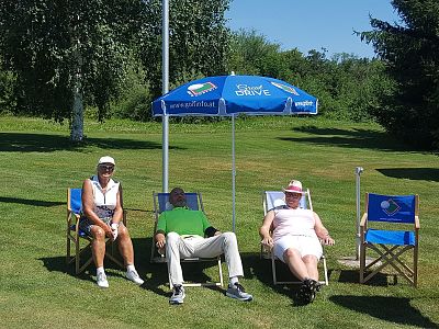 Drei Personen sitzen entspannt auf Liegestühlen unter einem Sonnenschirm auf einem grünen Rasen. Zwei Frauen und ein Mann genießen das sonnige Wetter.