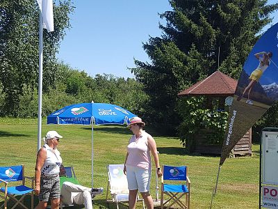 Zwei Frauen stehen auf einem Golfplatz neben Sonnenschirmen und Stühlen. Im Hintergrund sind Bäume und ein Holzgebäude zu sehen. Es ist ein sonniger Tag.
