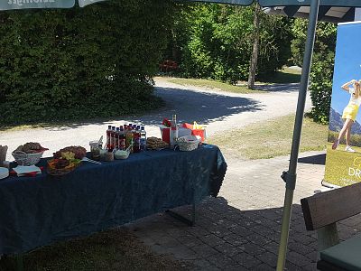 Buffet-Tisch im Freien mit verschiedenen Getränken und Snacks unter einem Sonnenschirm. Ein Plakat steht daneben, umgeben von einer grünen Landschaft.