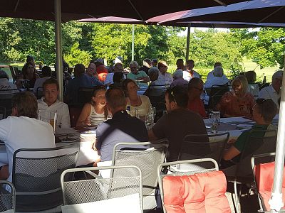 Menschen sitzen unter Sonnenschirmen in einem Restaurant im Freien. Sie genießen Mahlzeiten an mehreren Tischen bei sonnigem Wetter und grüner Umgebung.
