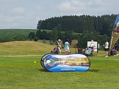 Golf course scene with players and a promotional banner in the foreground, displaying travel imagery. Trees and green hills are in the background.
