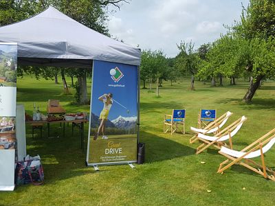 A golf event setup with a canopy tent, promotional banners, and wooden deck chairs on a grassy field with trees in the background.