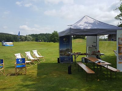 A promotional tent set up on grass with deck chairs, banners, a canopy tent, and picnic benches on a sunny day.