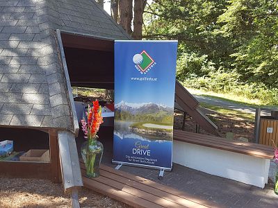 A rustic wooden shelter with a golf event banner and vibrant flowers, set in a wooded area with sunlight filtering through trees.