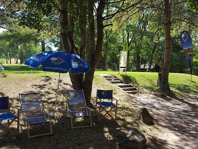 Foldable chairs under a blue umbrella in a sunny park with trees and greenery.