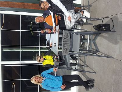 Four people sitting at an outdoor café table, smiling and enjoying their drinks. They are dressed in casual and sporty outfits on a sunny day.