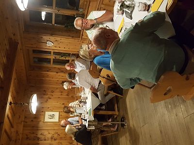 A group of people sitting around two tables in a wooden cabin with warm lighting, engaged in conversation and enjoying a meal. The setting is cozy and informal.