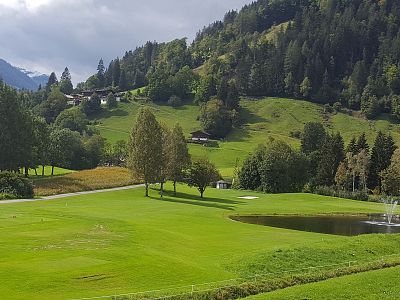 A picturesque golf course with green fairways, a pond with a fountain, and surrounding trees, set in a mountainous landscape under a clouded sky.