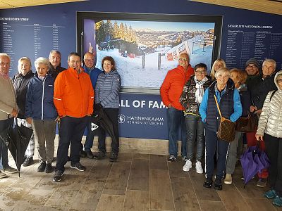 A group of 15 people in winter gear standing indoors by a ski resort display board. Some are holding umbrellas, suggesting they are prepared for diverse weather.