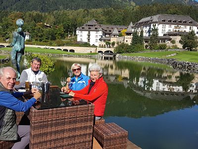 A group of four older adults sitting at an outdoor table by a lake with a scenic view of a large building and mountains in the background.