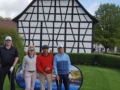 Four people in golf attire stand on grass before a half-timbered house, smiling, with a banner behind them and golf gear in hand.
