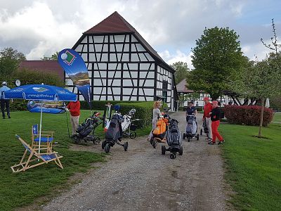 People with golf trolleys stand on a path near a half-timbered building, surrounded by green grass and trees, under a cloudy sky.