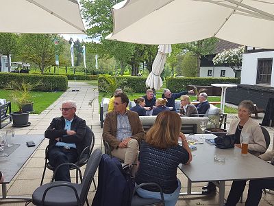 People seated at tables under umbrellas in a garden setting, engaging in conversation. Greenery and a building are in the background.