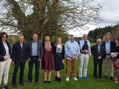 Gruppe von Menschen steht draußen auf einem Rasen vor einem Baum. Sie lächeln in die Kamera und tragen überwiegend legere Kleidung.