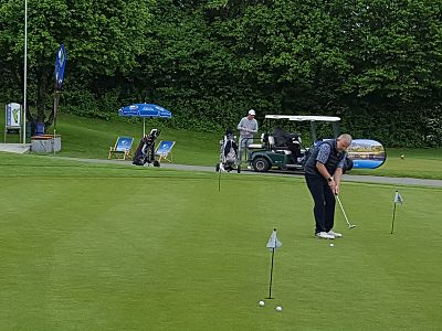 A golfer practices putting on a green with several golf carts and trees in the background on a cloudy day.