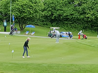 Golfers playing on a green course with trees, flags, and a golf cart in the background.