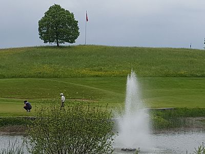 A golf course with a player putting near a fountain, trees in the background, and a cloudy sky.
