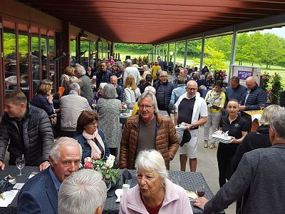 Crowd mingling at outdoor event under an awning, with tables and greenery.
