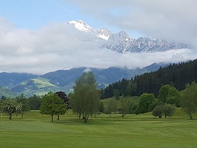 A lush green golf course in the foreground with trees, surrounded by a mountain range. Snowy peaks are visible under a blue sky with clouds.