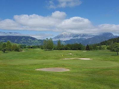 A green golf course with sand bunkers and mountains in the background under a blue sky with clouds.