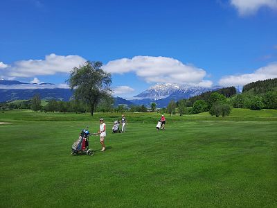 Golfers on a green course with mountains and blue sky in the background. Several players are visible with trees and distant hills under scattered clouds.
