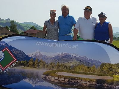Four people at a golf course in Austria stand behind a banner with a mountain and lake image on a sunny day.