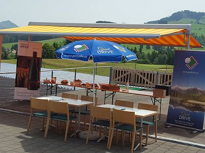 Outdoor event setup with a striped awning, banners, and a blue umbrella over tables on a sunny day, surrounded by a picturesque landscape.