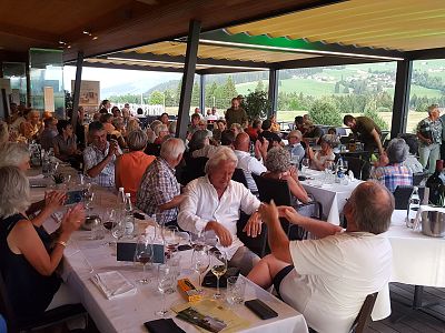 A group of people attending a wine tasting event indoors, with tables covered in white cloth, wine glasses, and bottles, surrounded by scenic view.