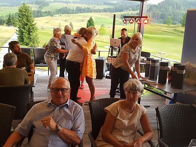 A group of people socializing at a golf club, with some hugging and others sitting at tables, set against a backdrop of green fields and hills.