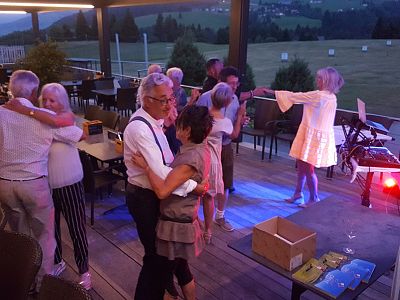 A group of people dance and socialize on an outdoor terrace at night, with scenic views in the background. The mood is lively and joyful.
